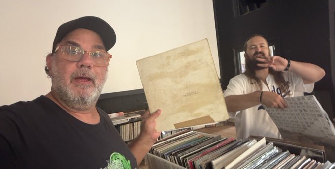 Two men pose with vinyl records; one wears a black cap and t-shirt while holding an empty record sleeve, and the other in a white shirt makes a peace sign.