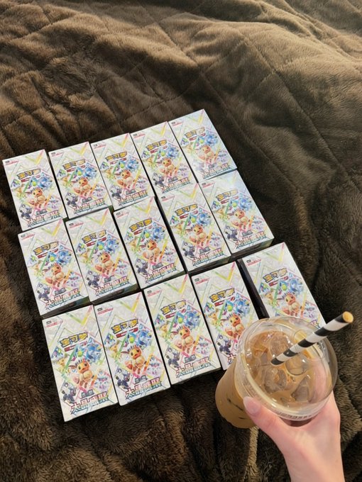 Multiple small boxed figures or merchandise arranged in rows on a brown blanket, with a hand holding an iced coffee drink with a striped straw in the foreground.