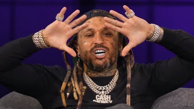 A muscular man with dreadlocks and tattoos, wearing a black shirt and multiple diamond rings and chains (one reading 'CASH'), posing with his hands framing his face against a purple background.