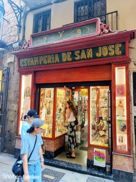 A storefront labeled "ESTAMPERIA DE SAN JOSE" in gold lettering on a red facade, displaying various religious prints and artwork in the windows, with a woman in a floral dress and a child in a grey cap standing outside.
