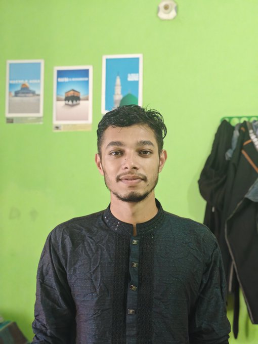 A young man with dark hair and a short beard wearing a black traditional button-up shirt poses for a portrait against a green wall decorated with small posters.