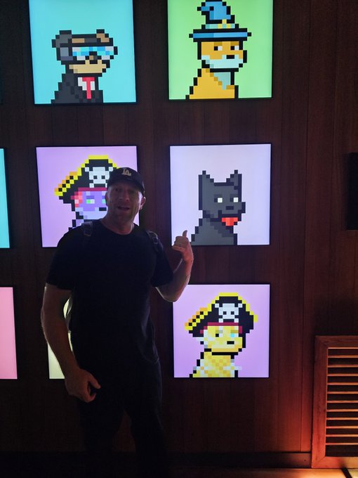 A man wearing a black cap and backpack gives a thumbs-up while standing in front of a display of illuminated pixel-art NFT dog portraits on wooden paneling.