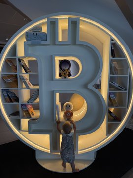 A child reaches up toward a large, illuminated Bitcoin logo designed as a bookshelf in a modern interior space.