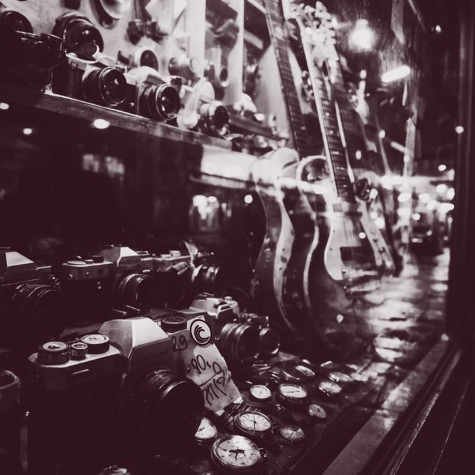 A black-and-white photograph of a shop window displaying vintage cameras, guitars, and pocket watches.