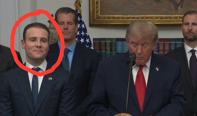 A man in a suit, circled in red, stands behind President Donald Trump as he speaks at a podium, likely at the White House. This likely relates to the post by referencing the man circled as someone who profited from an announcement.