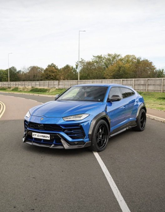 A blue Lamborghini Urus with carbon fiber accents parked on a road, displaying a "CT CARBON" front license plate.