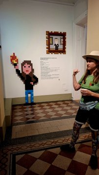 A woman wearing a beige hat, green t-shirt, and dark shorts stands next to a Super Mario pixel art display in what appears to be a museum or gallery exhibit.