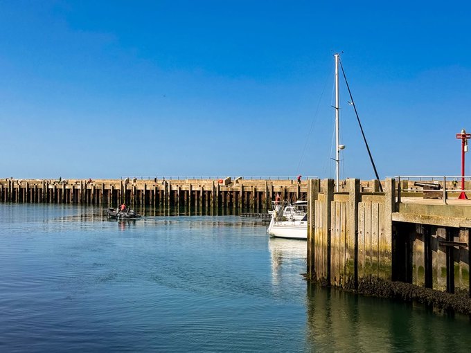 A scenic harbor view featuring a wooden pier extending into calm blue water, with a small sailboat moored alongside and people walking on the pier under a clear sky.