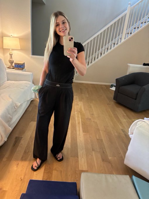 A young woman with long blonde hair takes a mirror selfie in a brightly lit living room, wearing a black short-sleeved top, black flowy pants, and black sandals, holding a phone with a beige case.