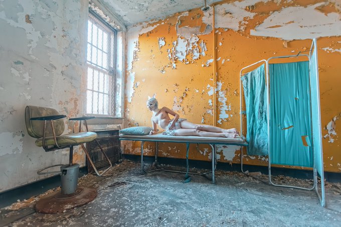 A photograph of a person in white lingerie, knee-high boots, and a mask reclining on a medical examination table within an abandoned, decaying room featuring peeling yellow paint and a privacy screen.