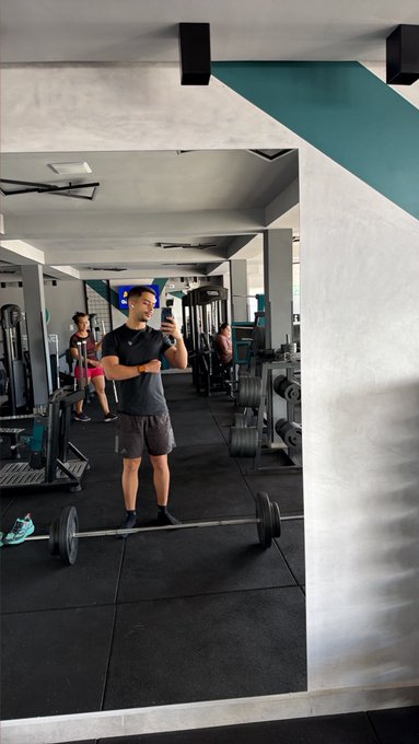 A gym selfie in a mirror showing a man standing with crossed arms behind a barbell on the floor, with gym equipment and other people in the background.