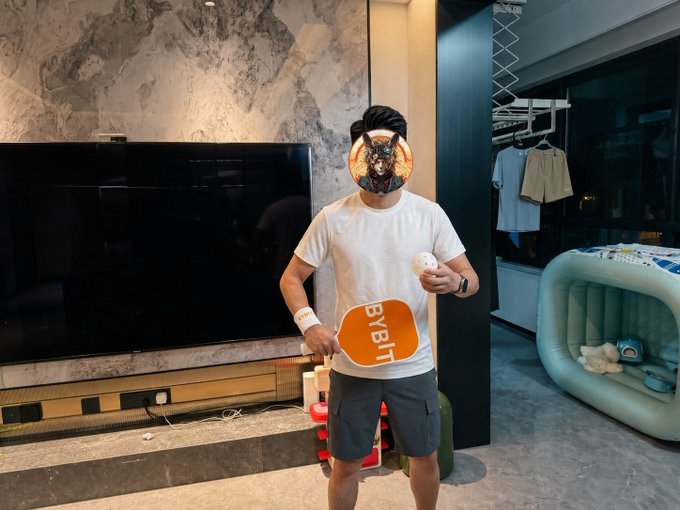 A man stands in a modern living room wearing a white t-shirt, gray shorts, and a dog-shaped face mask, holding a pickleball paddle branded with "Bybit" in one hand and a ball in the other. The pickleball kit's orange and white color scheme playfully matches his casual outfit, as referenced in the caption.