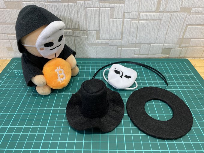 A plush figure in a black hoodie and white mask sits on a green cutting mat holding a Bitcoin coin, next to a disassembled black hat/cape costume piece and matching mask accessories.