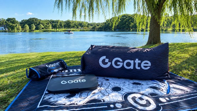 A picnic blanket, inflatable sofa, and small table branded with the "Gate" logo are arranged on grass beside a tranquil lake with a willow tree, matching the post's description of a Mid-Autumn Festival gift box from Gate Exchange that's perfect for camping. The accompanying text celebrates Gate's milestone of surpassing 60 million global users with a 127% reserve ratio.