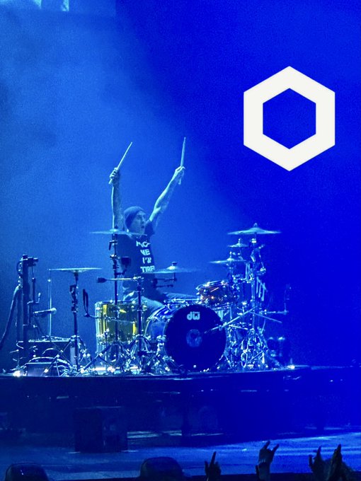 A drummer on stage with arms raised holding drumsticks triumphantly, with a large white hexagonal logo displayed on the blue-lit screen behind them.