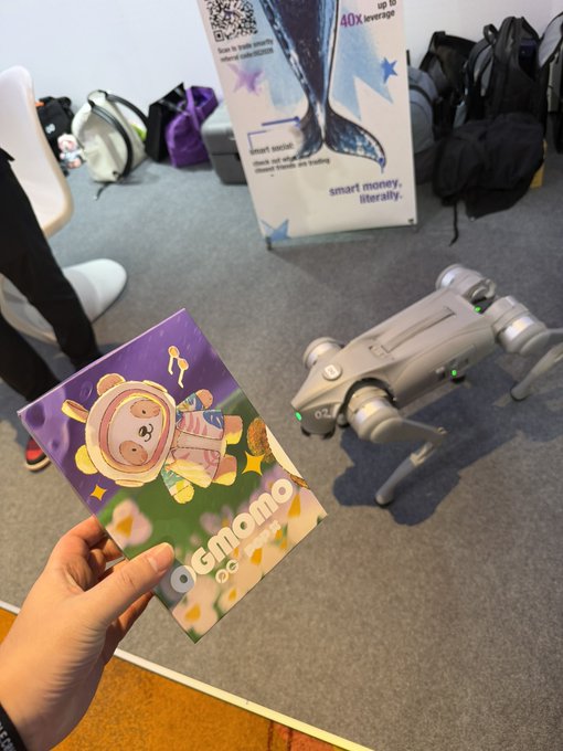 A photo taken at an indoor event where a person holds a colorful 'ogmomo' brochure next to a white quadruped robot dog, with a sign reading 'smart money, literally' in the background.