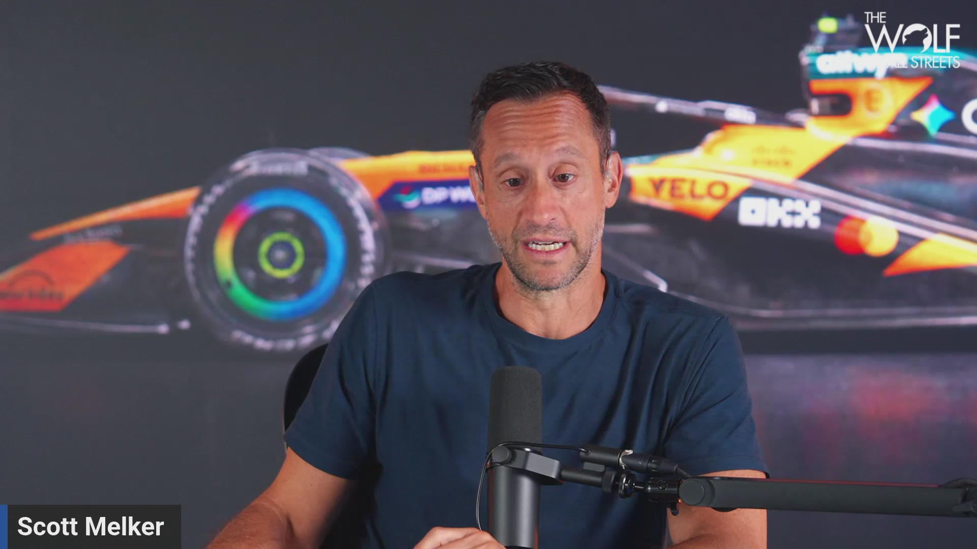 The image features a man sitting behind a microphone, wearing a blue shirt. He appears to be talking about Bitcoin or discussing its value. The background of the image shows an advertisement for a car race featuring a yellow car with a black stripe on the side. The text in the image reads "Bitcoin PLUMMETS Below $62K As Arthur Hayes DUMPS Zcash". This suggests that the man might be discussing the cryptocurrency market or Bitcoin, which is often associated with high-profile events and achievements.