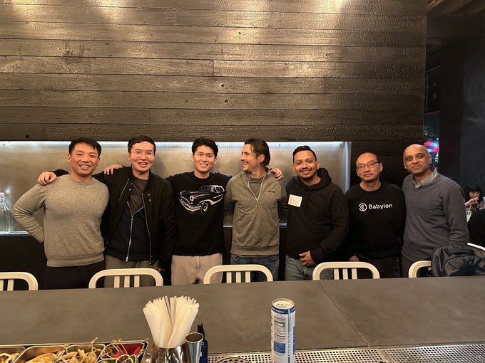 Seven men pose together smiling at what appears to be a casual social gathering in a wood-paneled venue, with one wearing a "Babylon" branded sweatshirt. The group photo, featuring founders and investors from crypto venture firms, aligns with the post's caption celebrating genuine industry networking moments in 2026.