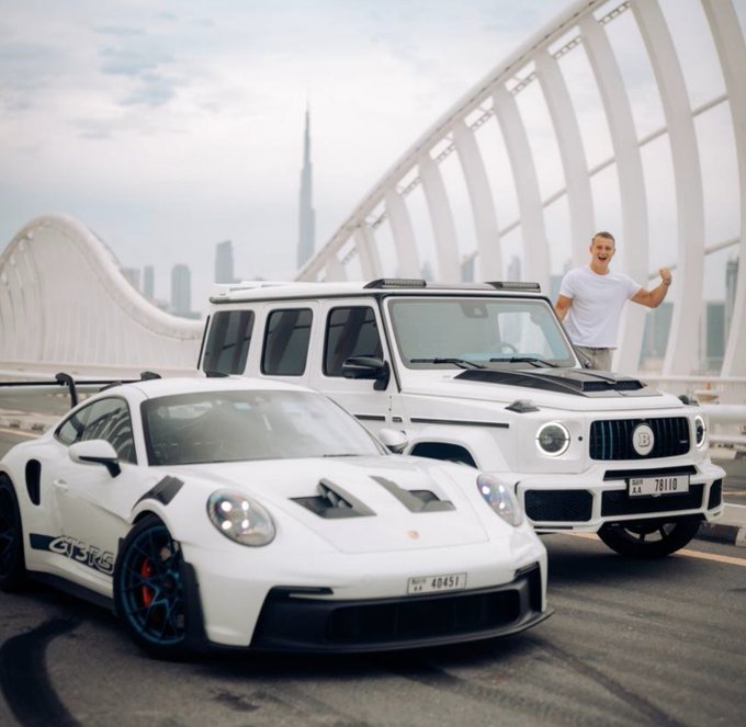 A smiling man in a white t-shirt stands triumphantly beside a white Porsche GT3 RS and a white Mercedes-Benz G-Wagon on a bridge, with Dubai's skyline (featuring the Burj Khalifa) in the background. The post suggests this photo symbolizes his financial transformation from being broke to immense wealth, achieved through Bitcoin and altcoin trading success.