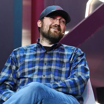 A bearded man wearing a baseball cap, glasses, and a blue plaid shirt smiles while sitting on a panel stage.