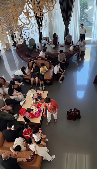 An overhead view of a large group of people gathered in an elegant lounge with ornate chandeliers, sitting on sofas and chairs around coffee tables laden with snacks and gifts.