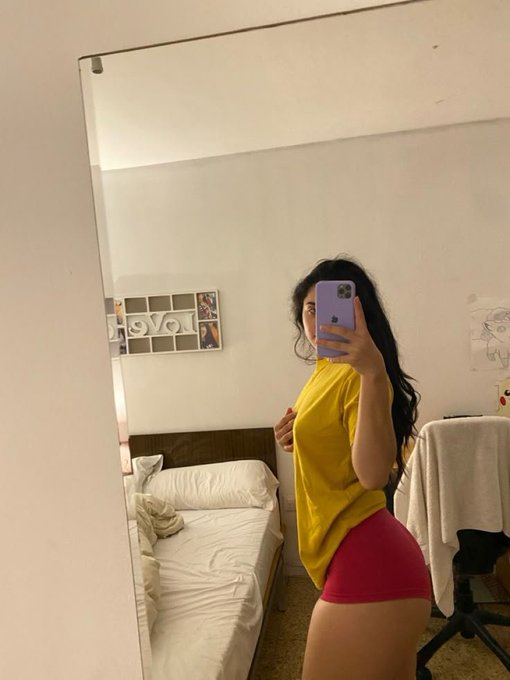 A woman in a yellow shirt and pink shorts takes a mirror selfie with her phone in her bedroom, which features a bed, wall decorations including the letters "LOVE," and an office chair. The "Gm" (good morning) caption pairs with the casual morning mirror selfie vibe.