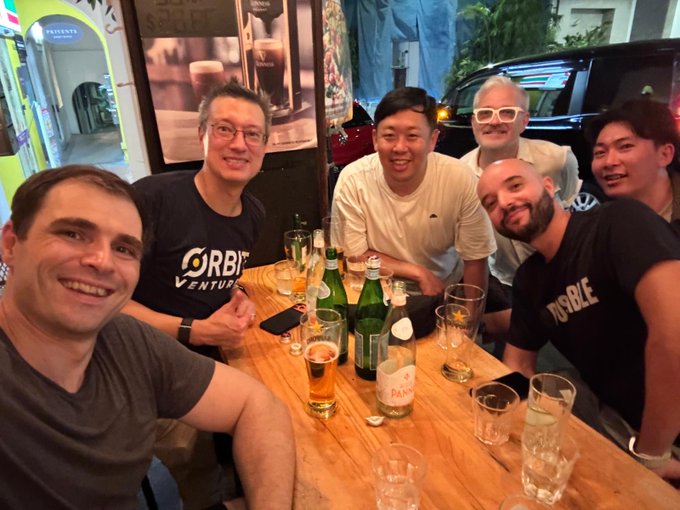 Six men are posing for a group photo at a wooden table in what appears to be a restaurant, with drinks, glasses, and bottles scattered across the table.
