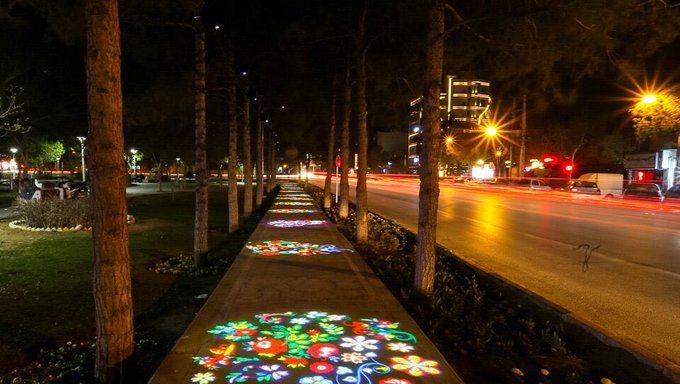 A nighttime scene of a tree-lined pedestrian walkway featuring colorful floral light projections on the pavement, with light trails from passing traffic on the adjacent road.