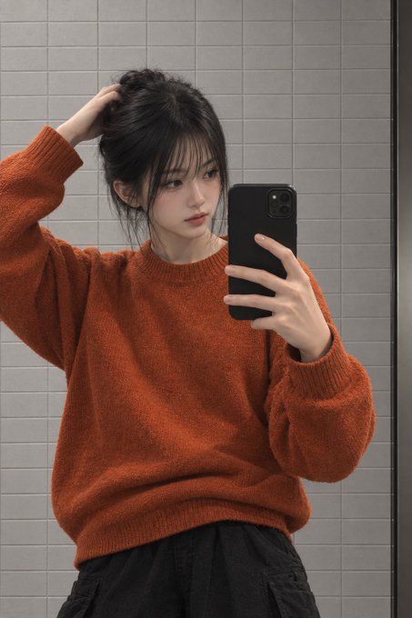 A young woman taking a mirror selfie with a smartphone, wearing an orange sweater and black pants, posing with one hand in her hair against a gray tiled wall.