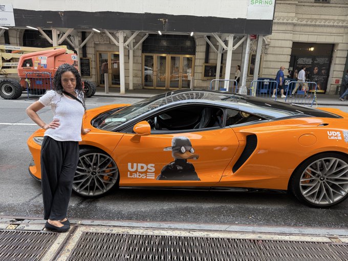 A woman stands next to an orange McLaren sports car featuring 'UDS Labs' branding and a cartoon character decal on its side.