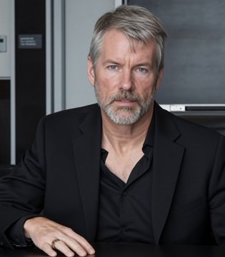 A portrait photograph of a distinguished older man with gray hair and a trimmed beard, wearing a black blazer over a dark shirt, seated at a table in a modern interior.