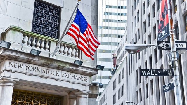 The New York Stock Exchange building is shown, prominently displaying the American flag and street signs for "Wall St," "Nassau," and "New," symbolizing the heart of U.S. financial markets. This iconic landmark directly illustrates the post's news that it is developing a 24/7 tokenized stock trading platform with on-chain settlement, exploring blockchain infrastructures like Avalanche.