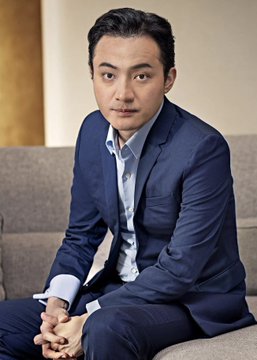 A man in a dark blue suit with a light blue shirt sits on a grey sofa, looking directly at the camera with his hands clasped.