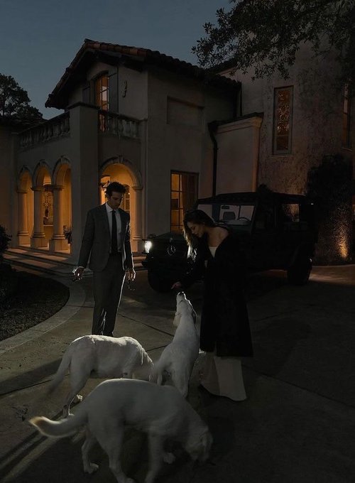 A man in a gray suit and woman in dark attire stand on a driveway at dusk, watching three white dogs play near a black SUV outside a Mediterranean-style home. The moody, dim lighting and carefree interaction with the dogs echo the post's sentiment of future prosperity, suggesting a glimpse into a lifestyle where present-day setbacks are overshadowed by the promise of success ahead.