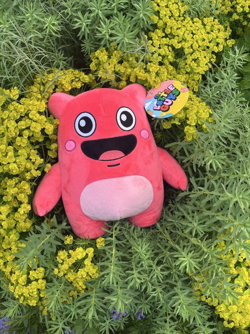 A red plush toy character with a big smile and white belly sitting among green foliage and small yellow flowers, with a colorful tag attached.