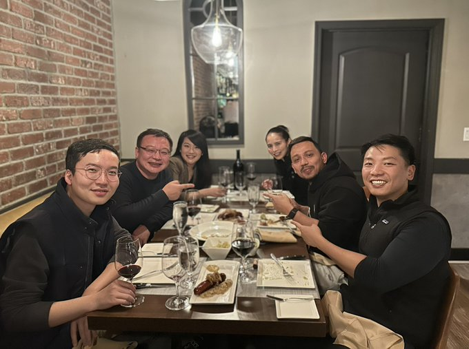 A group of six people enjoying a meal together at a restaurant table, smiling and posing for the camera with plates of food and wine glasses visible. The gathering reflects the celebratory and collaborative spirit behind the OKX @0xPolygon XL aggregation announcement in Palo Alto.