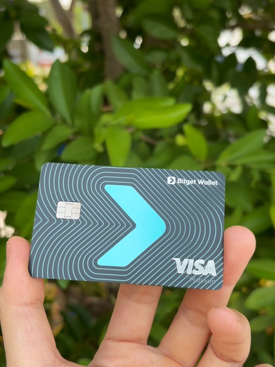 A hand holds up a gray Bitget Wallet Visa Platinum card featuring a striking turquoise chevron design against a backdrop of lush green foliage, visually showcasing the premium crypto-linked card mentioned in the post about earning up to 3% cashback in BTC, gold, stocks, or USDC.