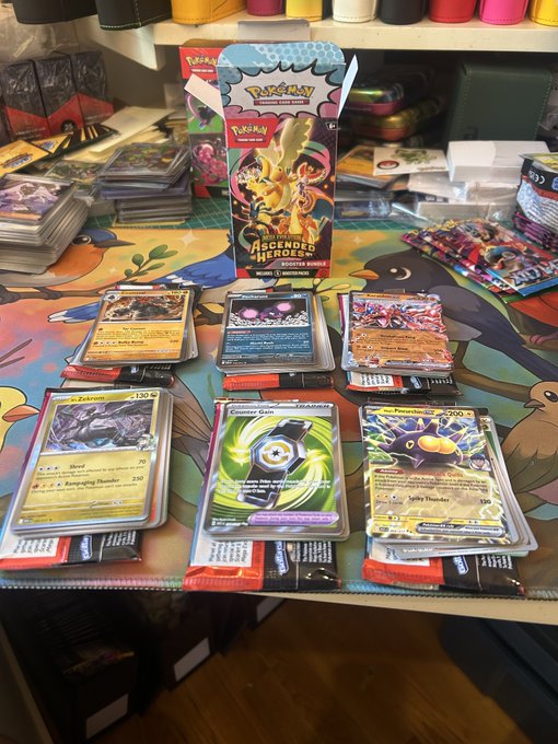 A collection of Pokémon trading cards arranged on a playmat, with a 'Scarlet & Violet Ascended Heroes' booster pack box in the background.