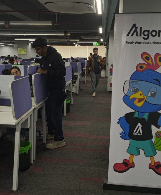An office workspace with rows of cubicles is shown alongside a tall banner for 'Algorand - Real-World Solutions' featuring a blue cartoon peacock mascot wearing a 'Bharat' shirt.