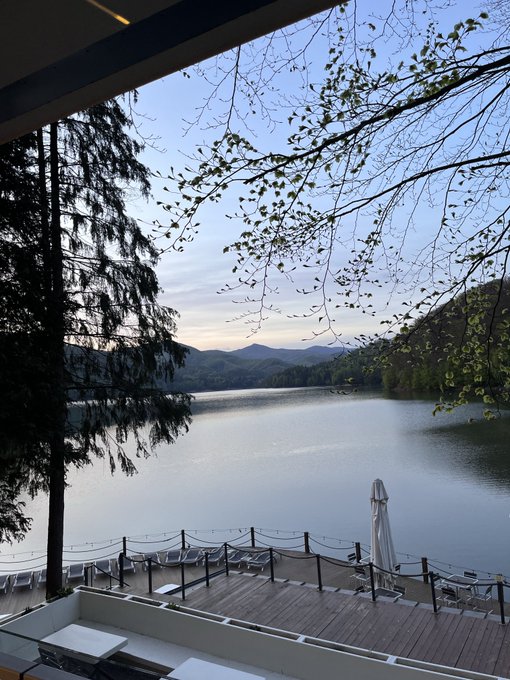 A serene view from a balcony overlooking a calm lake surrounded by forested mountains, with lounge chairs and a closed white umbrella on the wooden deck.
