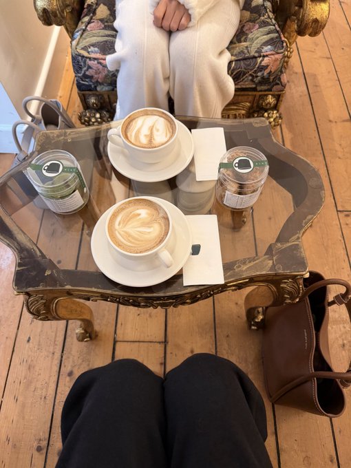 A view from above a glass coffee table holding two cups of latte art and two water bottles, with two people seated opposite each other on wooden floors.