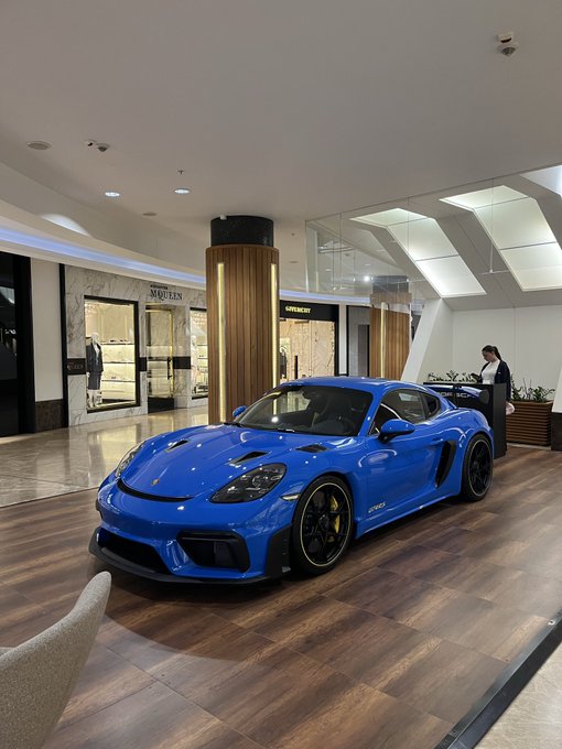 A blue Porsche GT2 RS sports car displayed in a luxury showroom with McCrea and Aiomersent boutiques visible in the background.