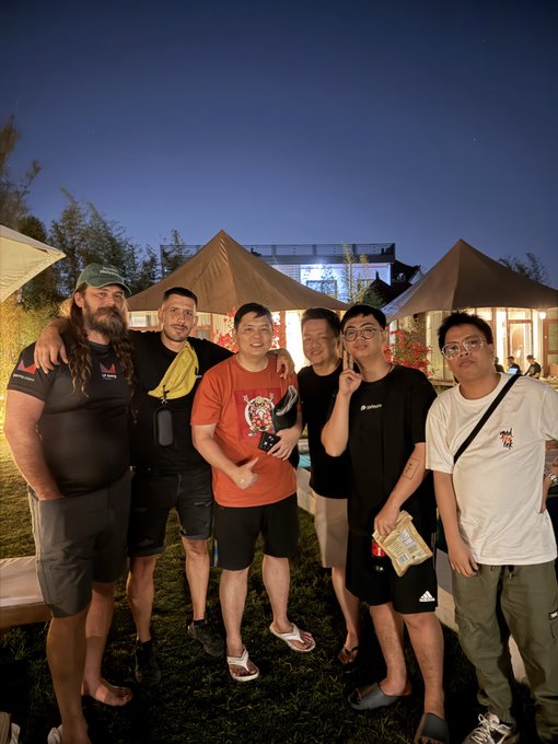 Six casually dressed men pose together on a grassy outdoor venue at dusk, with white tents and an illuminated building visible behind them under a deep blue sky, capturing the friendly gathering between the poster and the Meta LPARMY team mentioned in the caption ahead of the MeteoraIDN event in Bali, Indonesia.