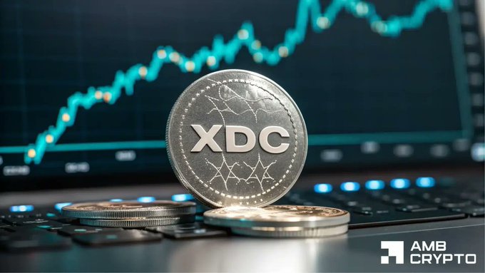 A silver physical XDC cryptocurrency coin standing on a keyboard with two other coins stacked beside it, set against a backdrop of a glowing cyan trading chart on a monitor, with an 'AMB Crypto' watermark.