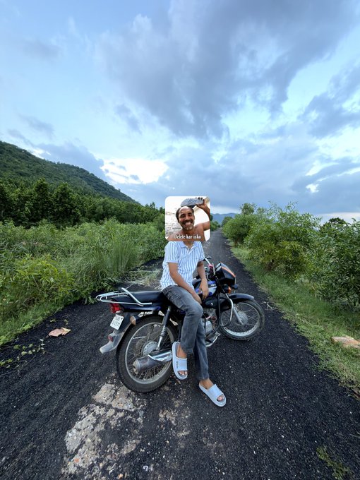 A man in a striped shirt and jeans sits on a black motorcycle on a rural road at dusk, with an overlay card reading 'Unter Karik' showing him taking a selfie.