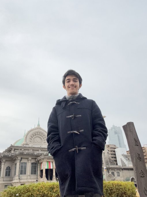 A young man in a black duffle coat smiles while standing outdoors with a historic building and city skyline in the background.