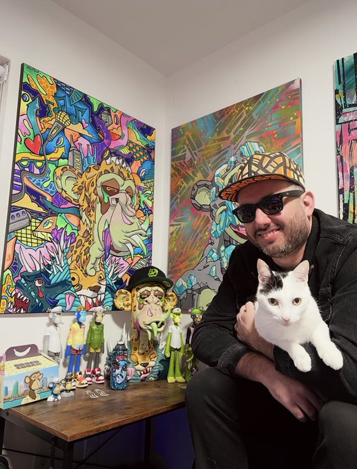 A man wearing a camouflage cap and sunglasses holds a white and black cat, sitting in front of colorful graffiti-style artwork and designer toys.