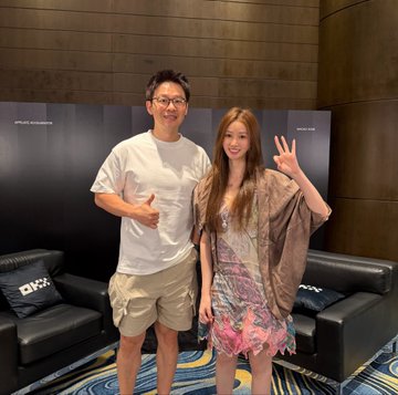 A man in a white t-shirt and glasses and a woman in a brown jacket and floral dress pose together in what appears to be a lounge area, both making peace signs.