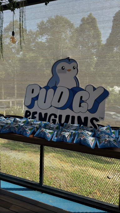 An outdoor scene featuring a 'Pudgy Penguins' sign with a cartoon penguin logo, placed behind a shelf filled with packaged snacks, with trees and a screened enclosure visible in the background.