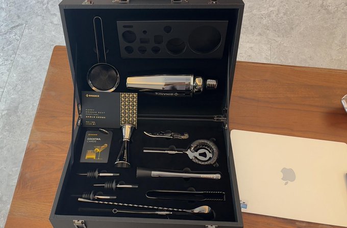 A premium black Binance VIP gift box is open on a wooden table beside a gold MacBook, revealing an assortment of engraved barware and cocktail tools—including a shaker, strainer, jigger, and stirrers—alongside a Cocktail Guide booklet and a members card, fitting the post's excitement about being upgraded to Binance VIP and receiving exclusive branded merchandise.